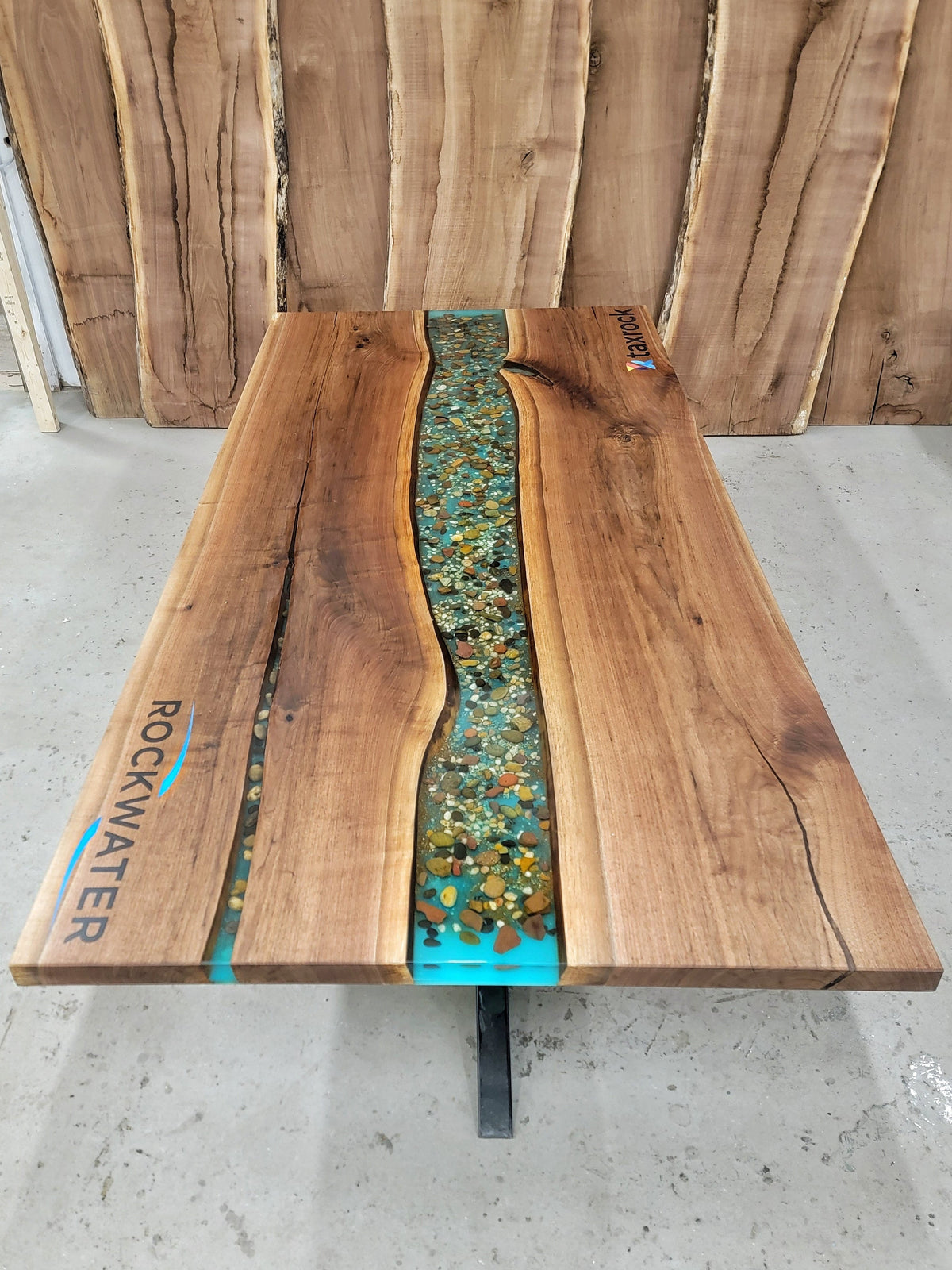 Walnut conference table with an embedded cntral epoxy river design filled with real stones, branded with engraved and epoxy-filled logo's that read 'Rockwater' and Taxrock', against a wooden panel background.