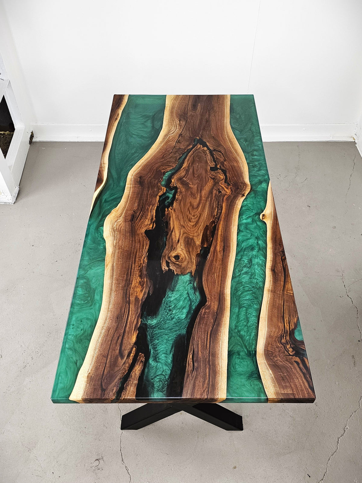 A live-edge wooden table with a vibrant green resin river design running through a natural wood grain, supported by a black metal X-frame base in a minimalist room.